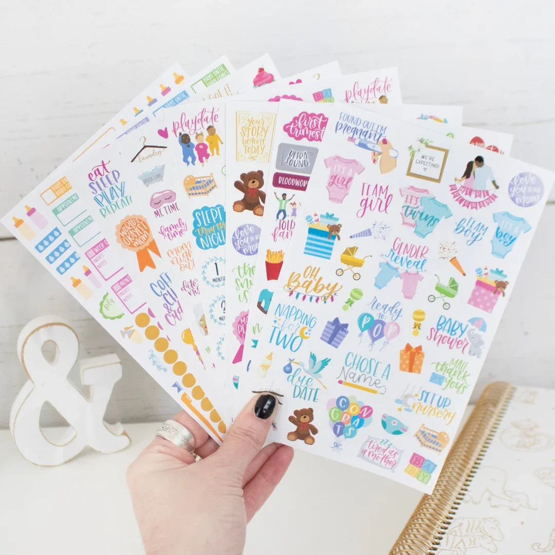 A hand holds bloomdailyplanners’ Planner Sticker Pack: Pregnancy & Baby’s First Year, featuring 5 colorful sheets with playful icons and phrases for baby milestones—ideal for memory keeping. A gold notebook and ampersand decor are in the background.