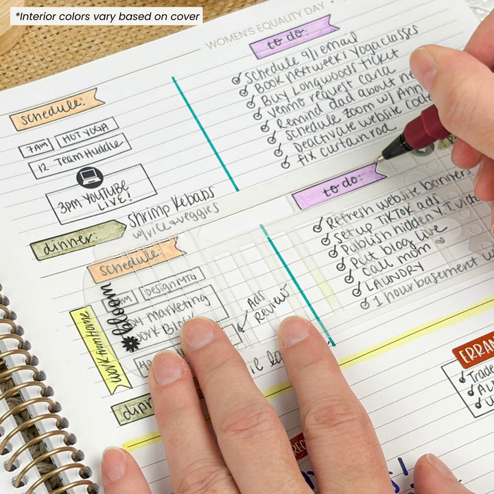 A close-up of a spiral-bound planner or notebook page with lined sections and colorful handwritten notes, with a hand holding a pen and another hand pointing at the page. A small text note in the top left says 'Interior colors vary based on cover.