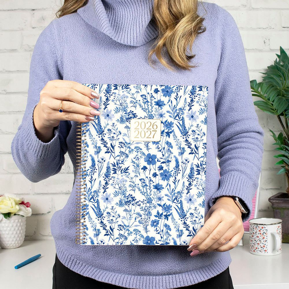 Person holding a floral-patterned planner with '2026-2027' on a white brick wall background.