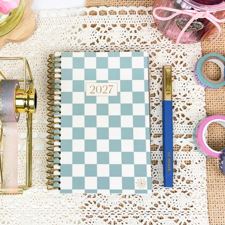 A spiral-bound planner with a light blue and white checkered cover and a small '2027' label, styled on a white lace runner with a blue pen, washi tape, and other desk accessories around it.