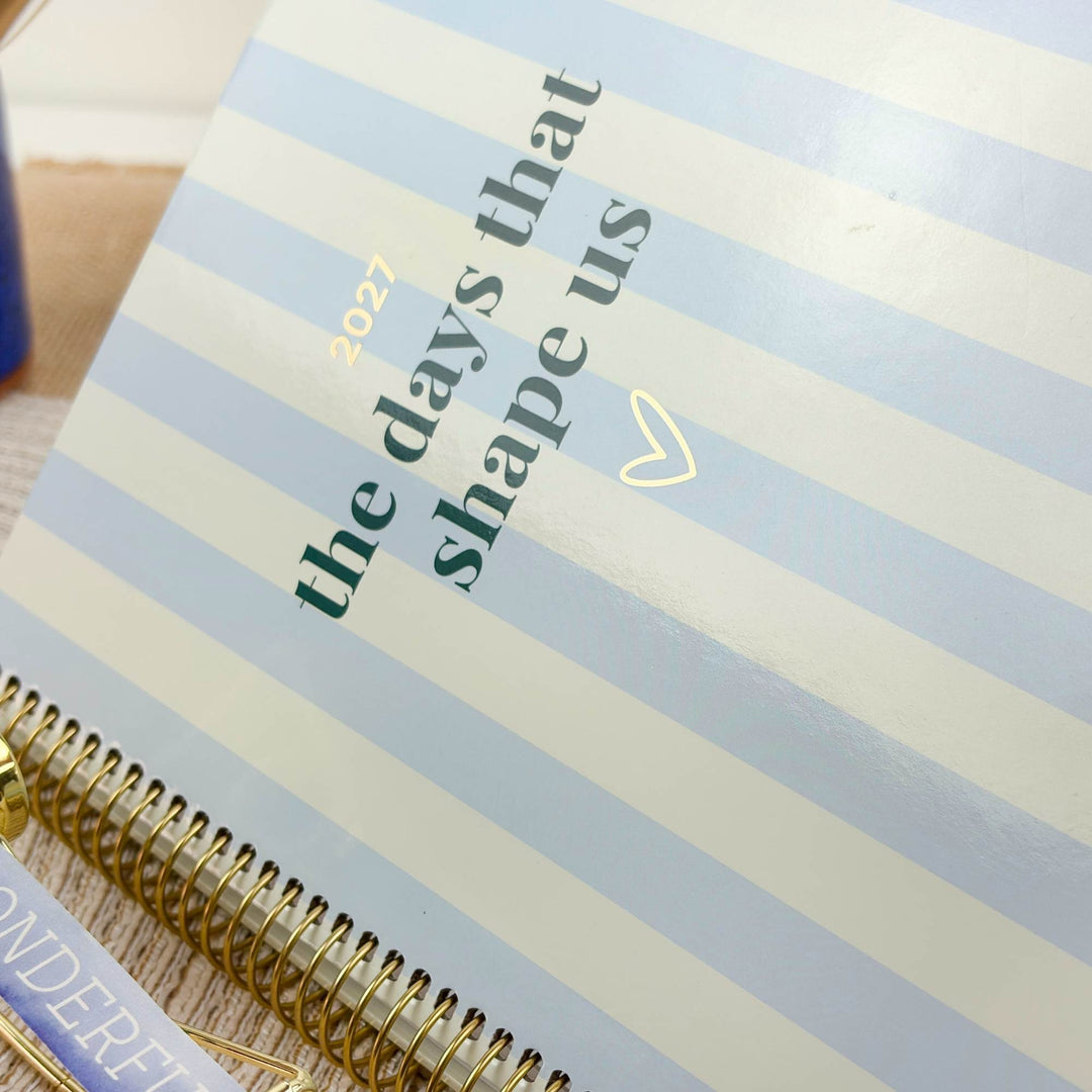 A close-up of a spiral-bound planner or notebook with pale blue and white horizontal stripes. The cover reads 'the days that shape us 2027' in dark green and gold text, with a small gold heart.