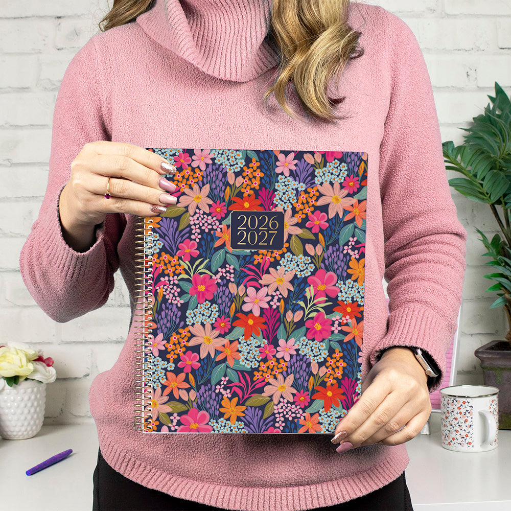 Person holding a floral planner with 2026-2027 dates, wearing a pink sweater.