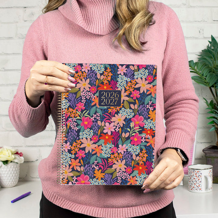 Person holding a floral planner with 2026-2027 dates, wearing a pink sweater.