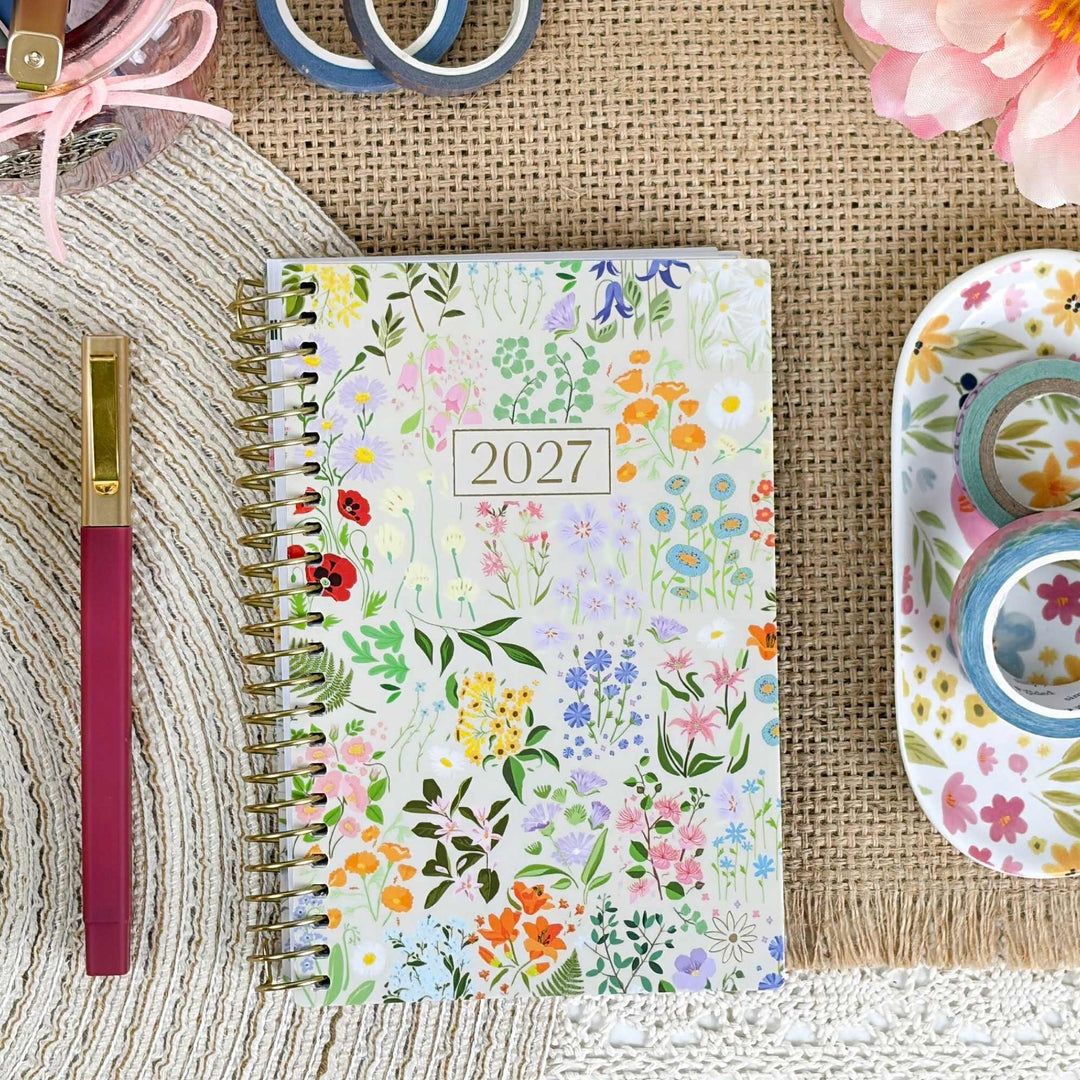 A 2027 spiral-bound planner with a colorful wildflower pattern, styled in a flat-lay arrangement with a pink pen, washi tape, and decorative paper on a burlap and knit background.