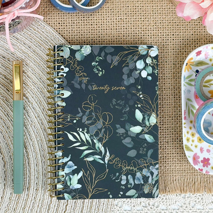 A spiral-bound planner or notebook with a dark floral cover lies on a woven mat, surrounded by stationery props including a mint pen, washi tape, and a small dish.