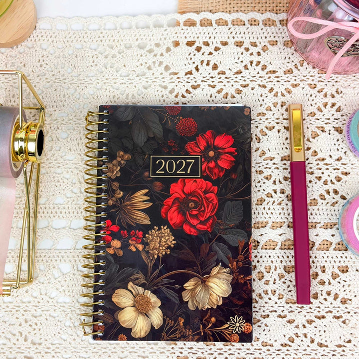 A 2027 spiral-bound planner with a dark floral cover featuring red and cream flowers, styled on a white lace doily next to a magenta and gold pen.