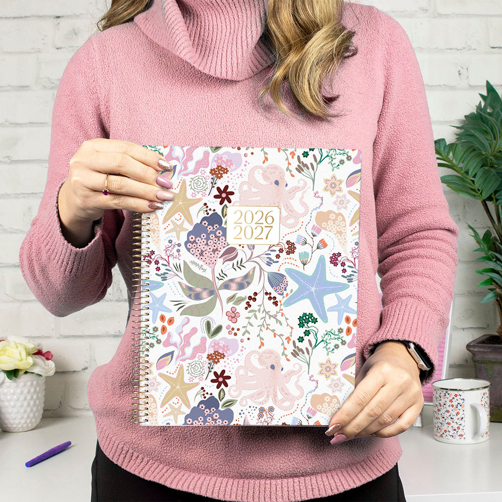 Person holding a floral planner with 2026-2027 dates, wearing a pink sweater.