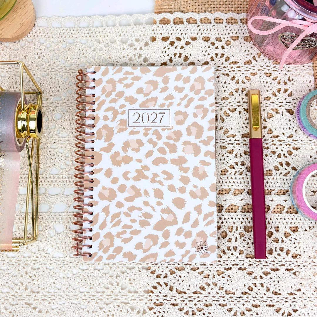 A spiral-bound 2027 planner with a beige and white leopard print cover, styled on a cream lace doily with a magenta pen, washi tape, and stationery accessories.