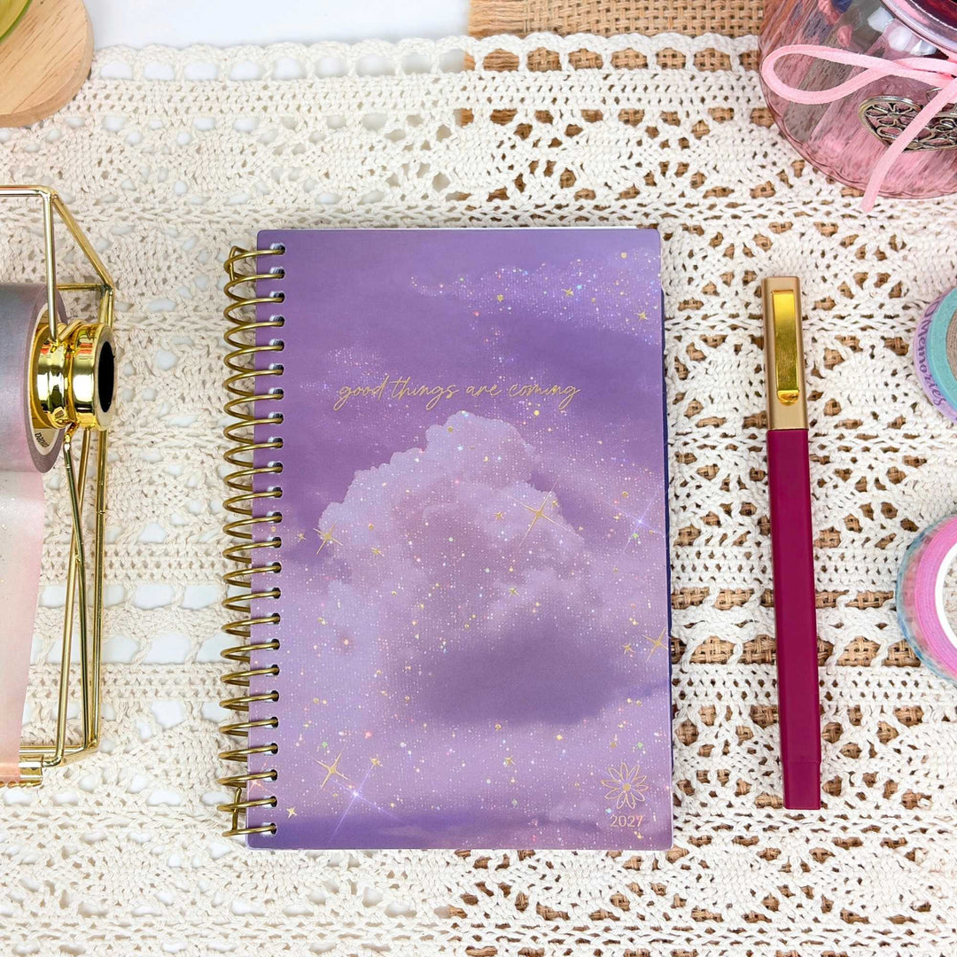 A spiral-bound planner or notebook with a lavender and purple cloud design and gold glitter accents, placed on a white lace surface next to a magenta and gold pen.