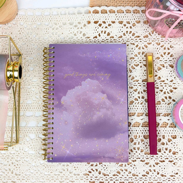 A spiral-bound planner or notebook with a lavender and purple cloud design and gold glitter accents, placed on a white lace surface next to a magenta and gold pen.