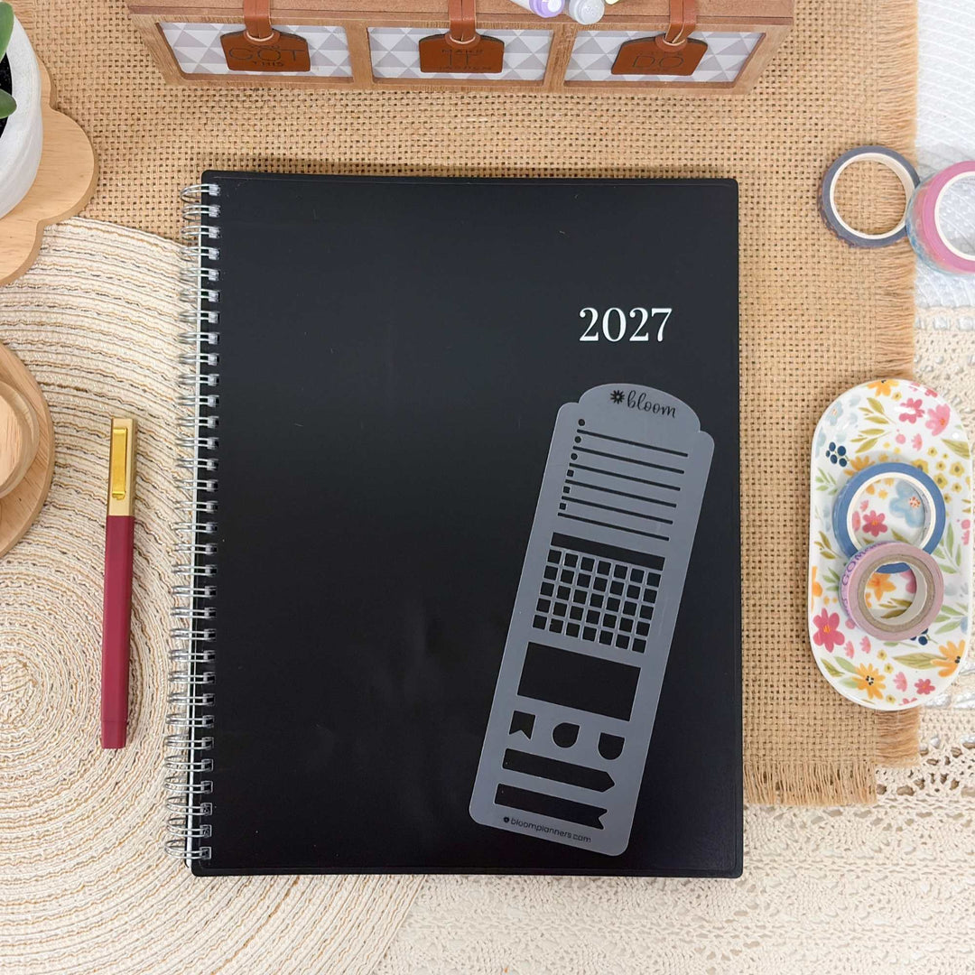 A black spiral-bound planner or notebook with the year 2027 and a gray calendar-style graphic on the cover, styled on a woven beige mat with stationery props around it.