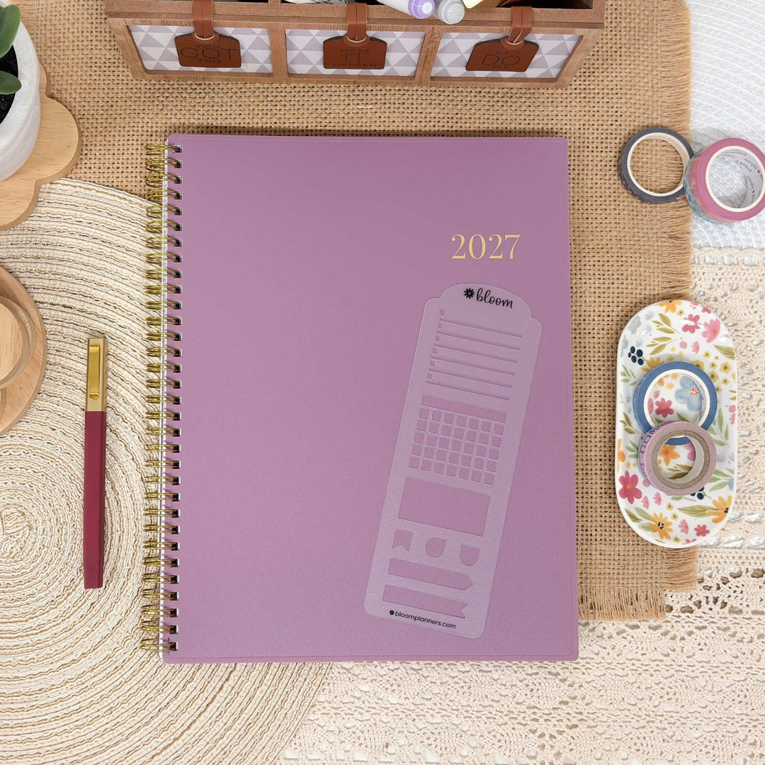 A 2027 spiral-bound planner with a dusty purple cover and gold foil details, styled in a flat-lay arrangement with washi tape, a paintbrush, and other desk accessories.