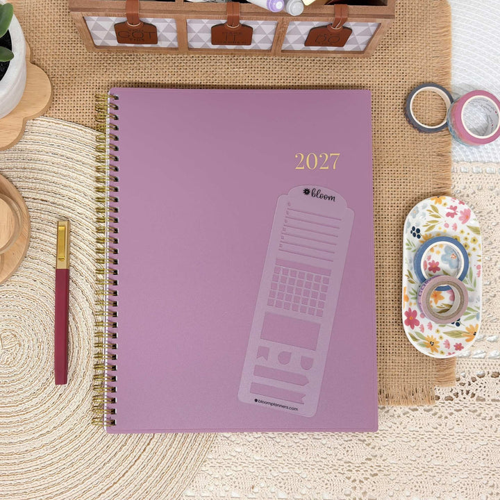 A 2027 spiral-bound planner with a dusty purple cover and gold foil details, styled in a flat-lay arrangement with washi tape, a paintbrush, and other desk accessories.