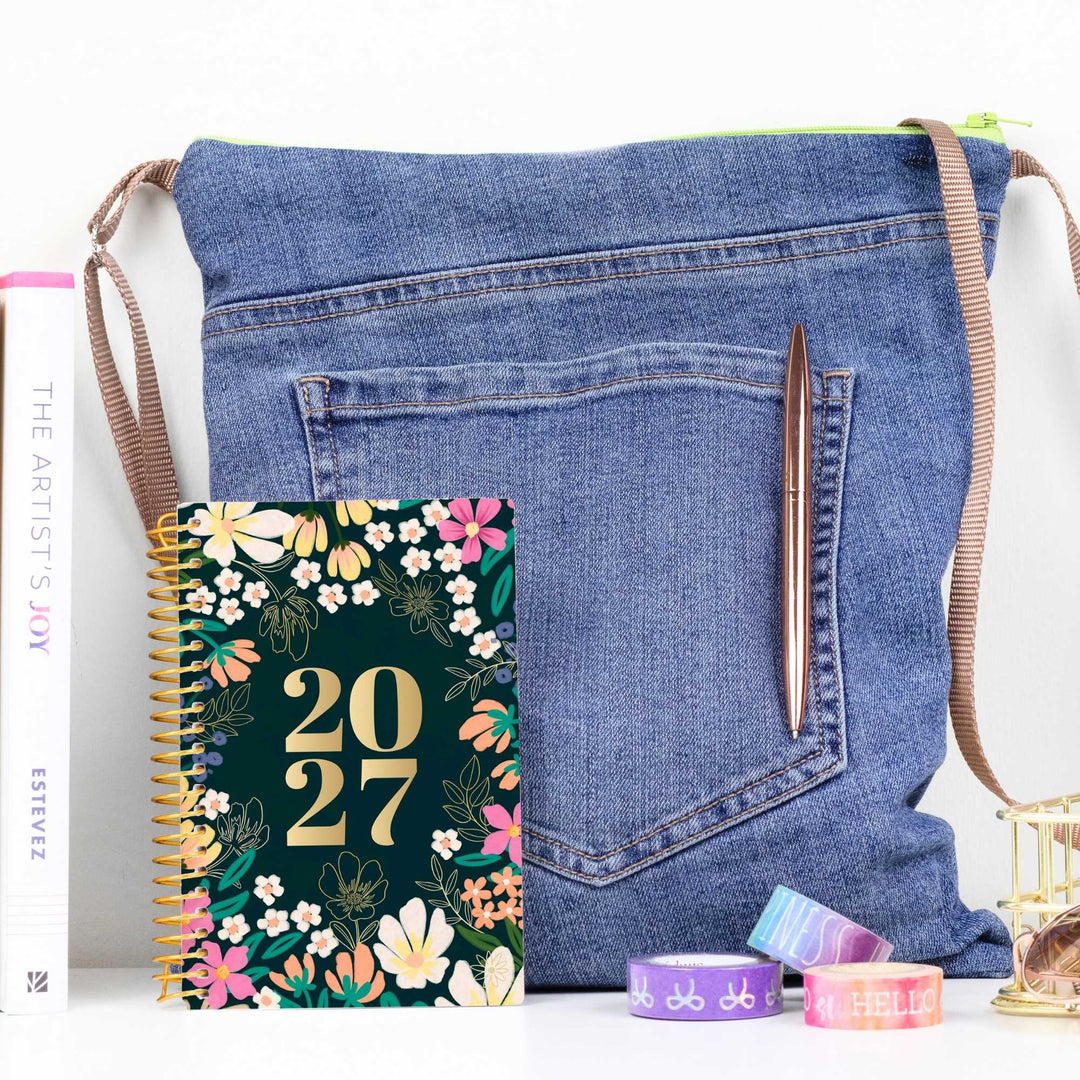 A denim shoulder bag styled with a spiral planner, a pen, washi tape, and a book on a white surface against a white background.