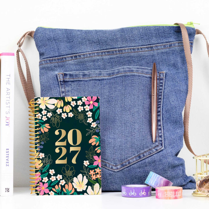 A denim shoulder bag styled with a spiral planner, a pen, washi tape, and a book on a white surface against a white background.