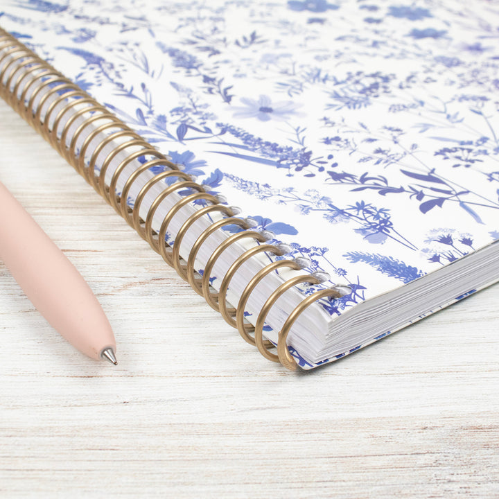 Planner with floral design on cover and gold spiral binding, accompanied by a pen on a light wooden surface.