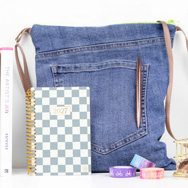 A blue denim shoulder bag styled with a leopard print 2027 planner, a brown pen, and colorful washi tapes on a white surface.