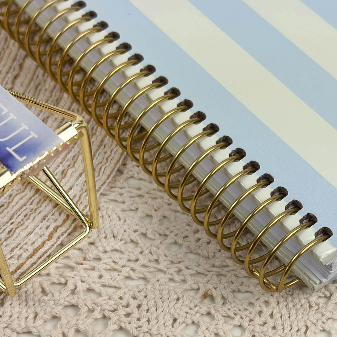 A close-up of a spiral-bound planner or notebook with a gold wire coil binding and a light blue and white striped cover, placed on a beige crocheted surface next to a gold geometric stand.