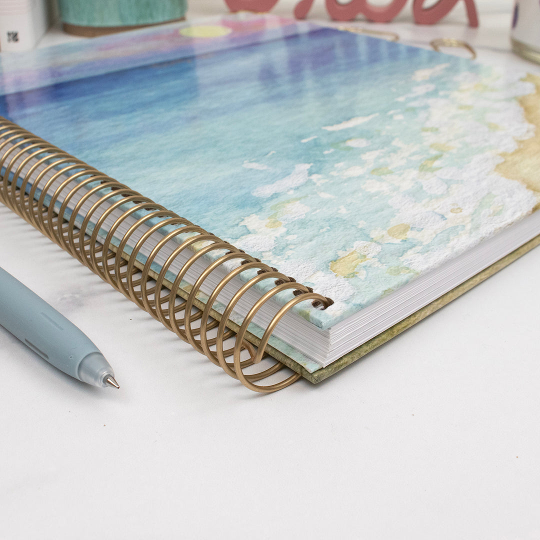 Planner with floral design on pages and gold spiral binding, accompanied by a pen on a white surface.