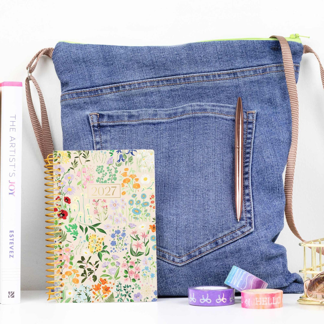 A blue denim crossbody bag styled with a floral 2027 spiral planner, a brown pen, and washi tapes on a white surface.