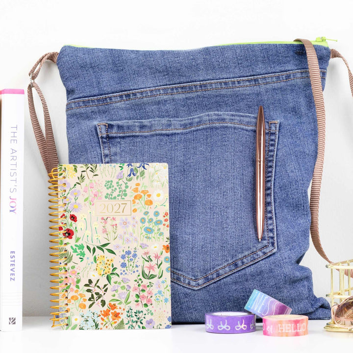 A blue denim crossbody bag styled with a floral 2027 spiral planner, a brown pen, and washi tapes on a white surface.