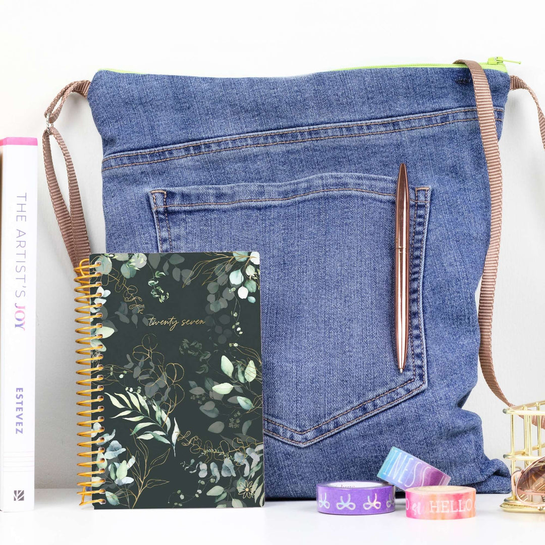 A blue denim shoulder bag styled with a spiral notebook, pen, and washi tape on a white surface.