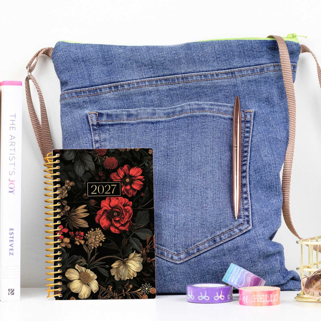 A blue denim shoulder bag styled with a floral 2027 spiral planner, a rose gold pen, and colorful washi tapes on a white surface.