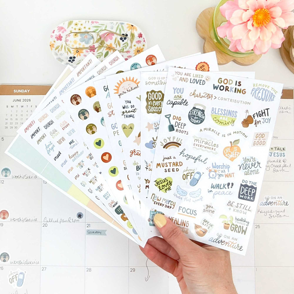 A hand holding fanned out sheets of Christian based planner stickers cver a planner on a desk. - hover