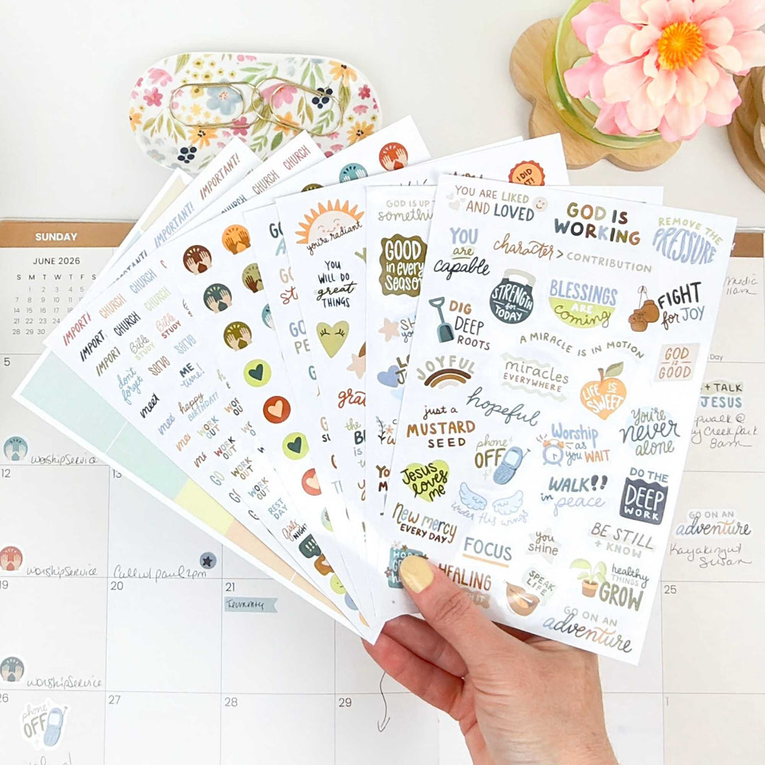 A hand holding fanned out sheets of Christian based planner stickers cver a planner on a desk.