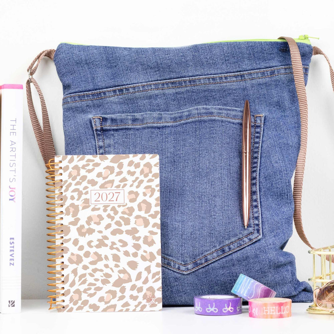 A blue denim shoulder bag styled with a leopard print 2027 planner, a brown pen, and colorful washi tapes on a white surface.