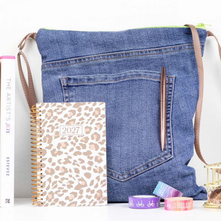 A blue denim shoulder bag styled with a leopard print 2027 planner, a brown pen, and colorful washi tapes on a white surface.