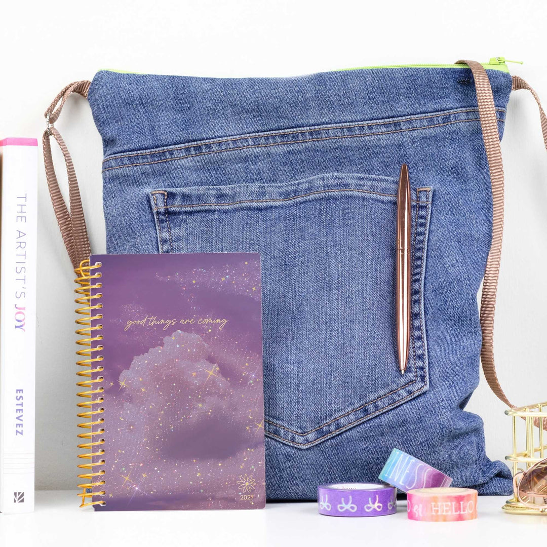 A blue denim shoulder bag styled with a purple spiral notebook, a brown pen, and colorful washi tapes on a white surface against a white background.