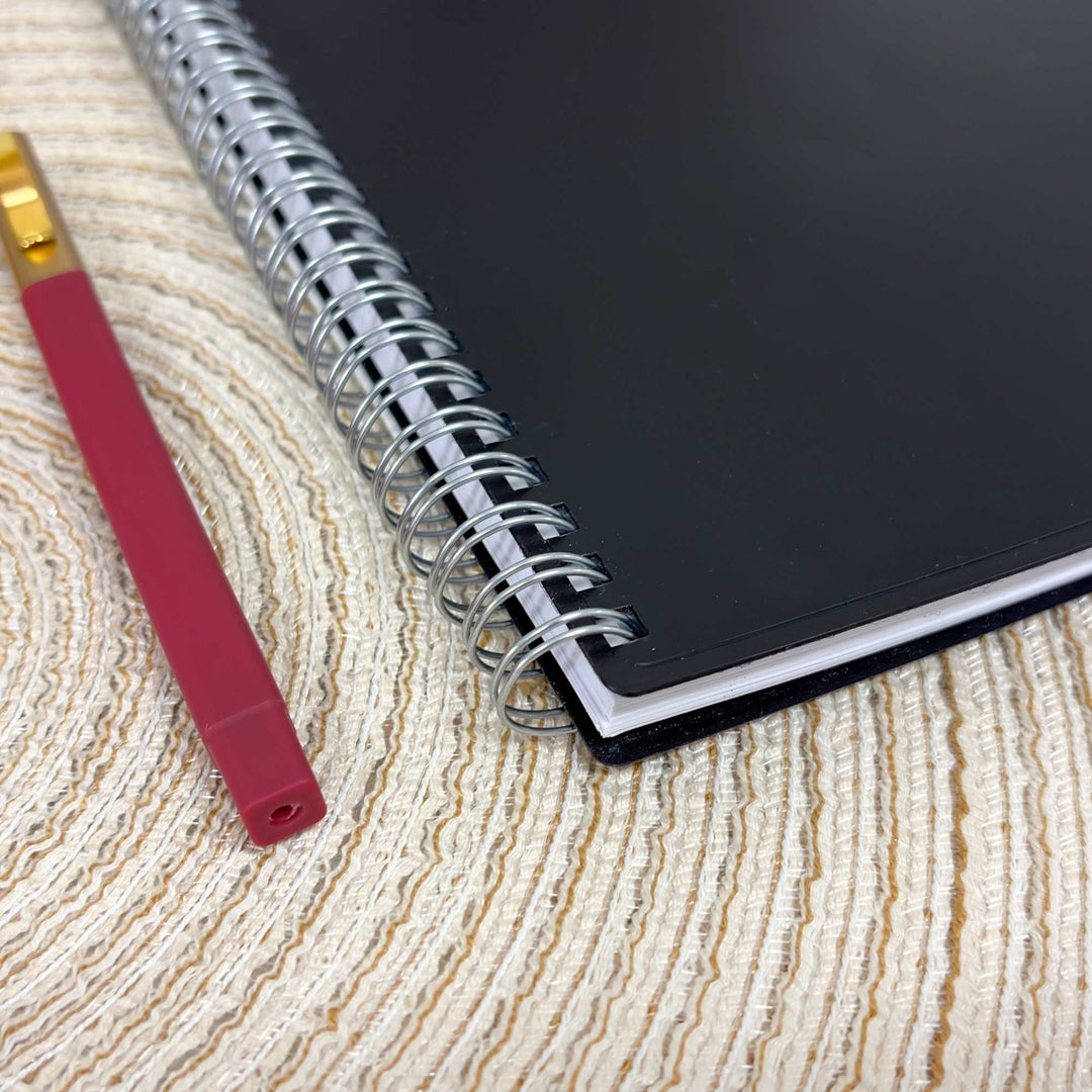 A black spiral notebook with a silver wire binding lies on a beige textured woven surface next to a red pen with a gold ferrule.