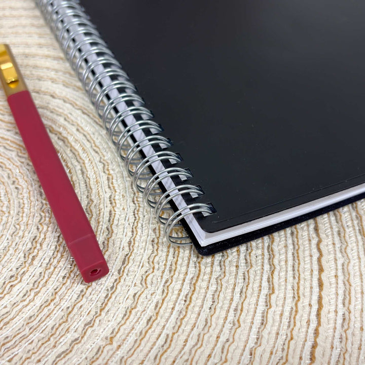 A black spiral notebook with a silver wire binding lies on a beige textured woven surface next to a red pen with a gold ferrule.