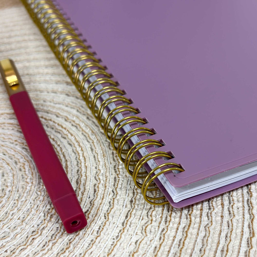 A purple spiral notebook with gold wire binding placed on a textured beige woven mat next to a dark red pen with gold accents.