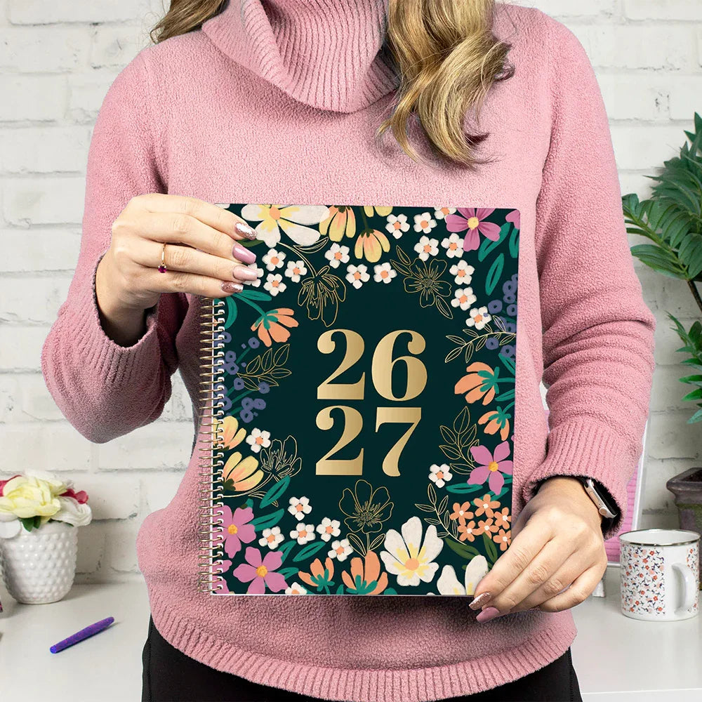 Person holding a notebook with floral design and '26 27' text, wearing a pink sweater.