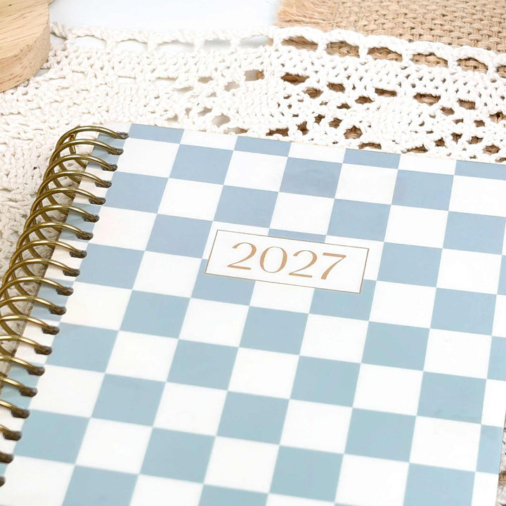 A spiral-bound 2027 planner with a light blue and white checkerboard cover, displayed on a white lace doily with a wooden surface in the background.