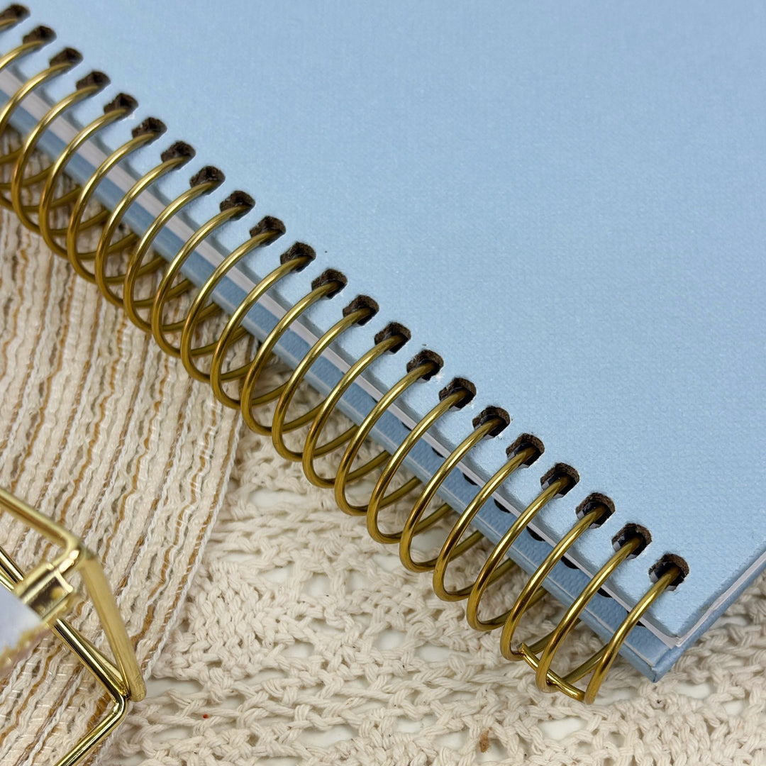 A close-up of a light blue spiral notebook with a shiny gold wire binding, resting on a textured beige lace fabric.