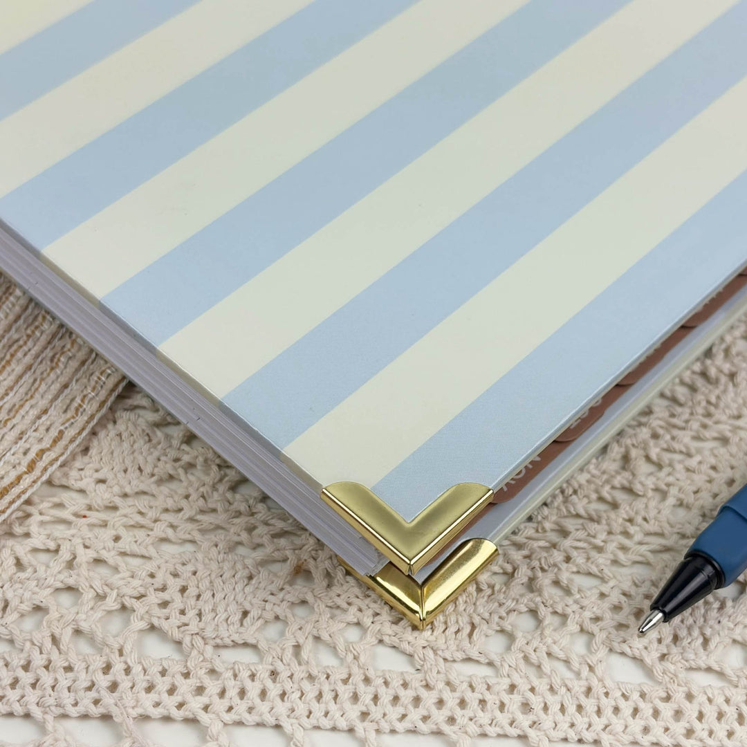 A close-up of a notebook or journal with pale blue and cream diagonal stripes, secured with a gold corner protector, resting on a beige crocheted surface with a blue pen partially visible.