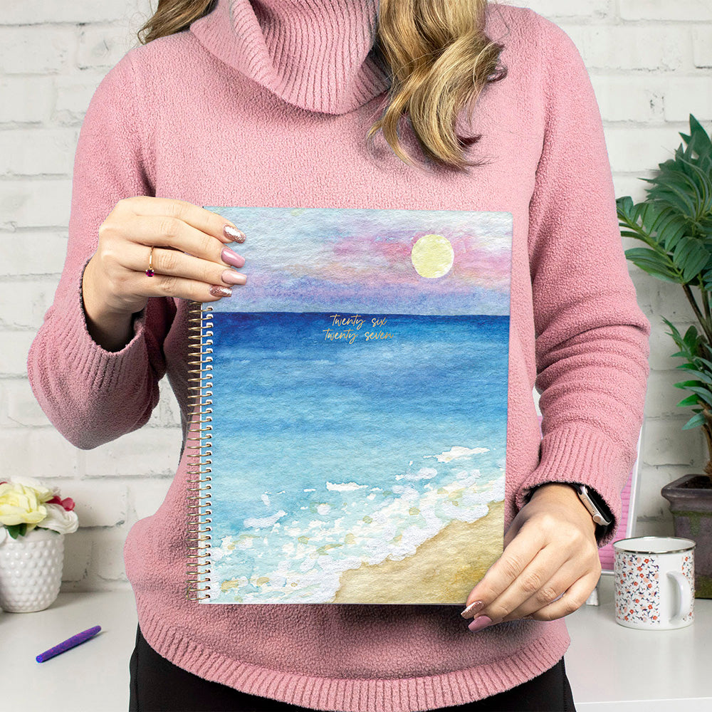 Person holding a spiral-bound notebook with a beach illustration, wearing a pink sweater.