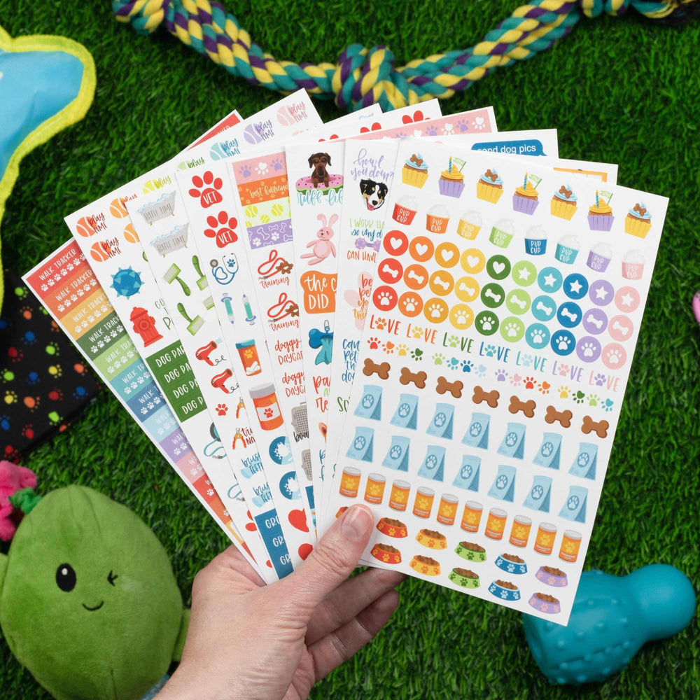 A hand displays bloomdailyplanners’ Planner Stickers, Dog Lover Pack—colorful, pet-themed designs like paw prints, bones, and cute sayings. Ideal for planners or journals. Pet toys rest on green grass in the background.