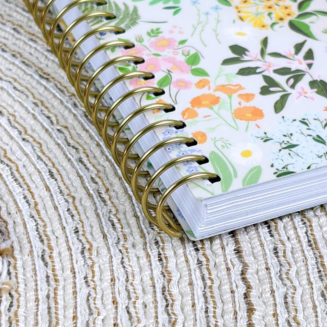 A close-up of a spiral-bound notebook with a white floral cover and shiny gold wire binding, resting on a textured beige woven surface.