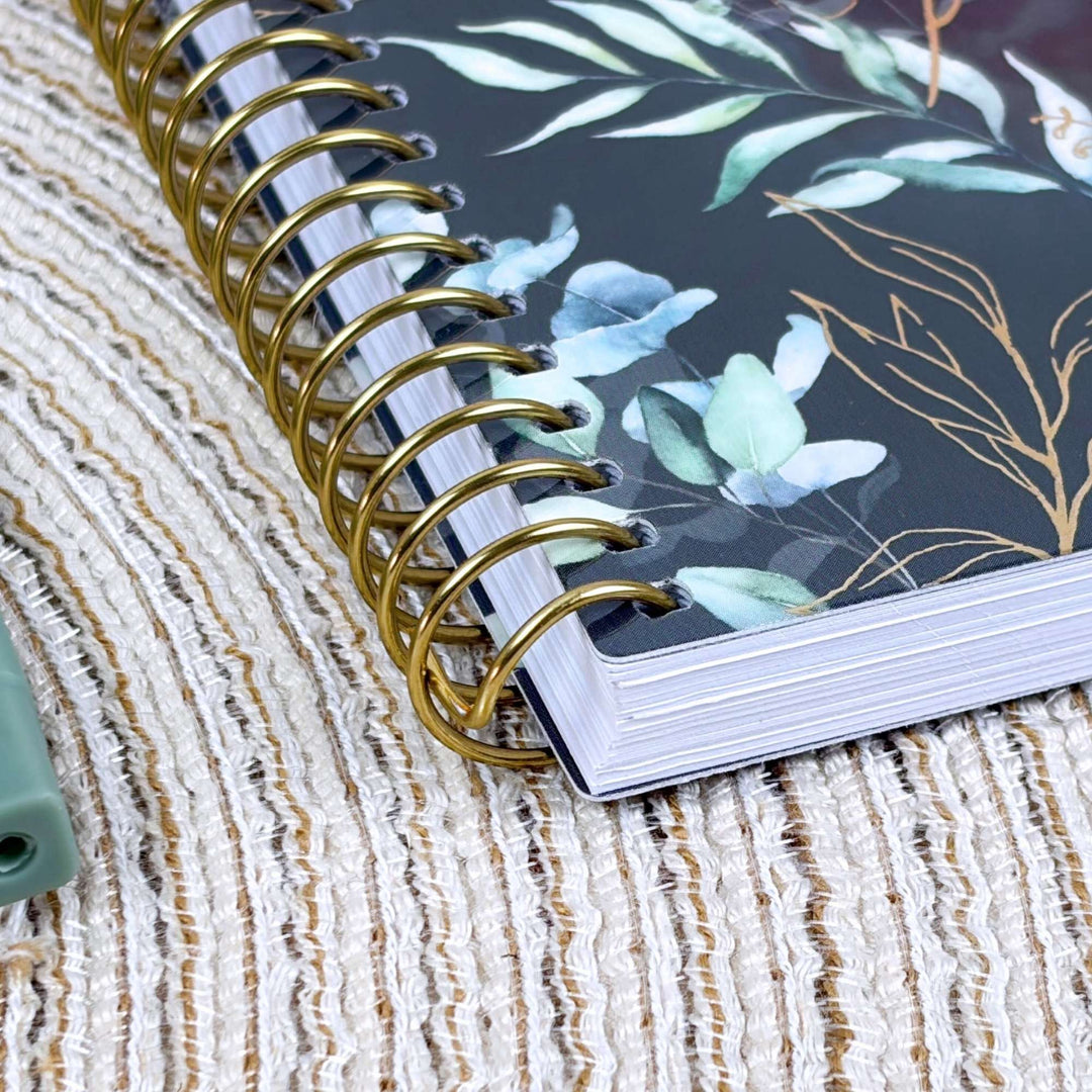 A close-up of a spiral-bound notebook with a black cover decorated with light blue and green botanical leaves and gold line art, resting on a textured beige woven surface.