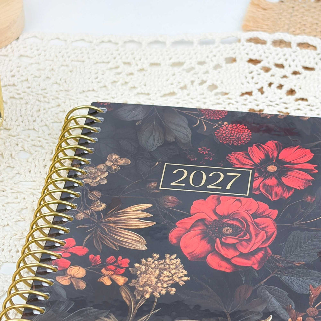 A 2027 spiral-bound planner with a dark floral cover, featuring red and pink flowers, gold leaves, and a gold spiral binding, styled on a white lace doily.