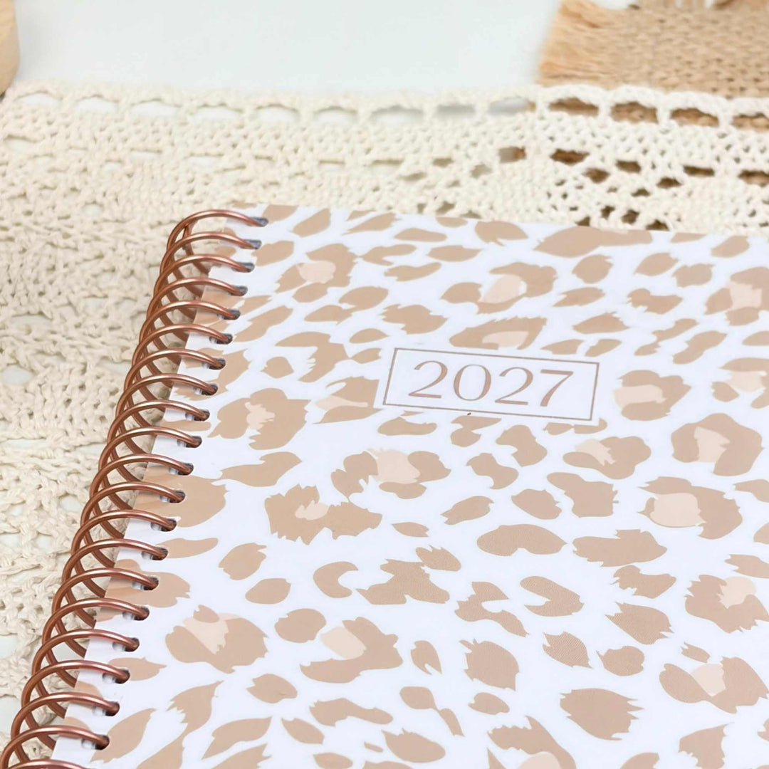 A spiral-bound 2027 planner with a white cover featuring a beige leopard print pattern, resting on a cream-colored crocheted lace surface.