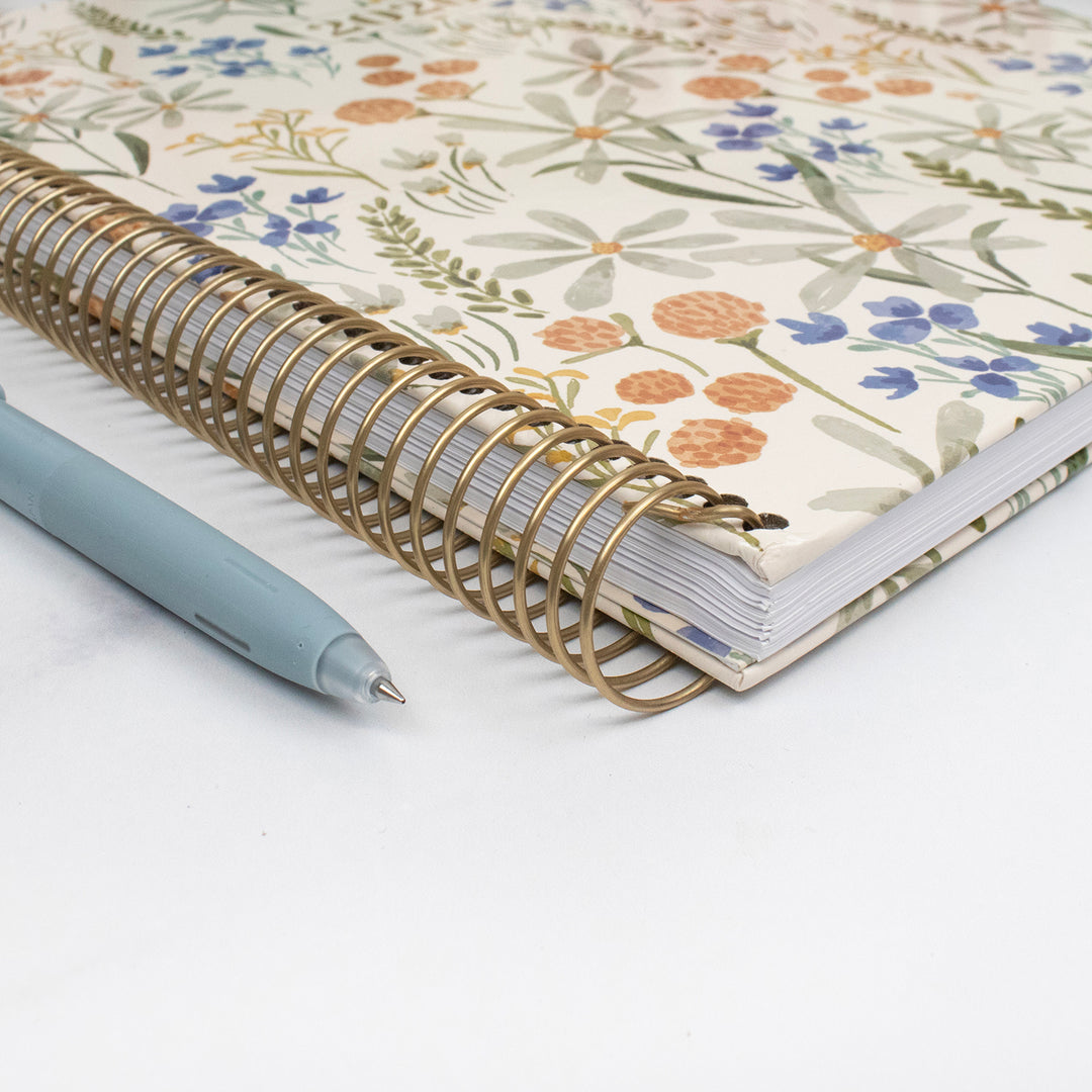 Planner with floral cover and gold spiral binding, accompanied by a pen on a white background