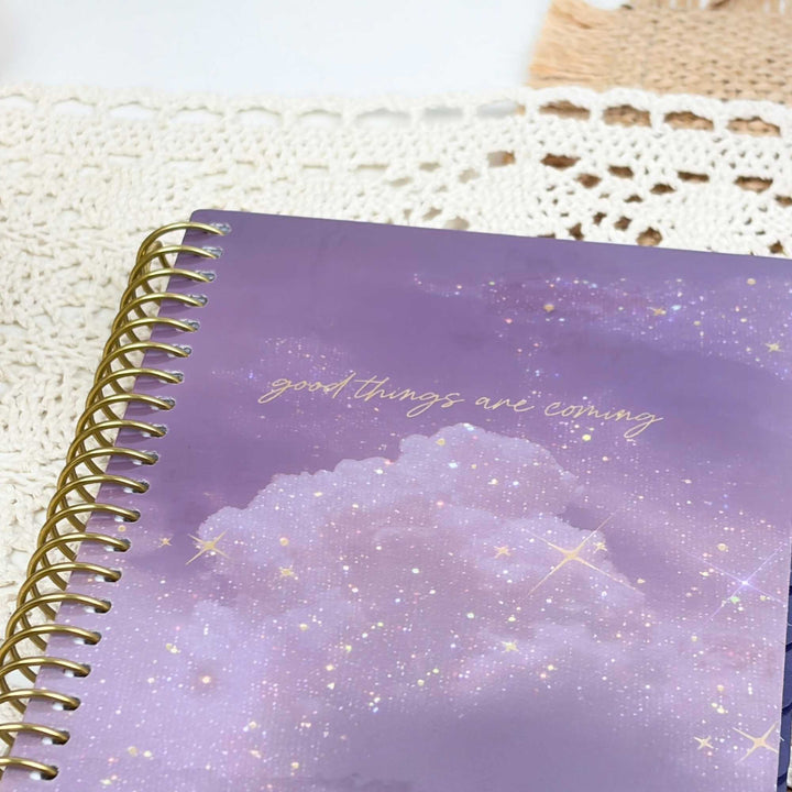 A spiral-bound notebook with a purple galaxy cloud cover and gold foil text reading 'good things are coming', photographed on a white lace doily.