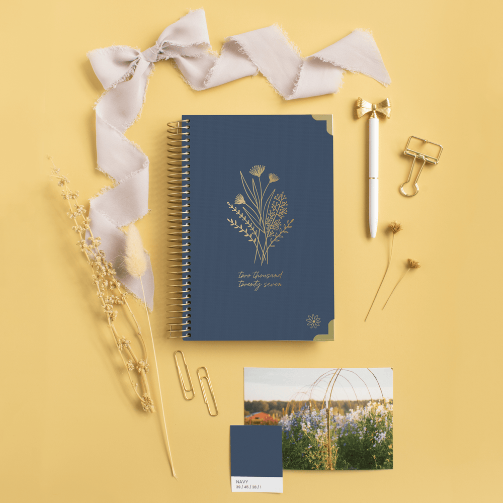 A navy blue spiral-bound planner with gold botanical line art and the words 'Two Small Things' is arranged on a warm yellow background with a pale pink ribbon, gold pen, binder clips, dried flowers, a color swatch card, and a small printed photo. - hover