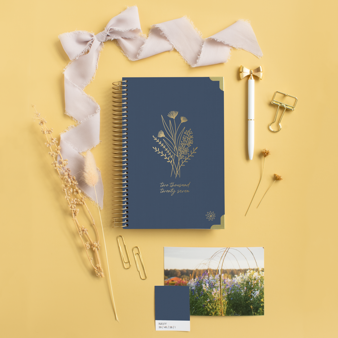 A navy blue spiral-bound planner with gold botanical line art and the words 'Two Small Things' is arranged on a warm yellow background with a pale pink ribbon, gold pen, binder clips, dried flowers, a color swatch card, and a small printed photo.