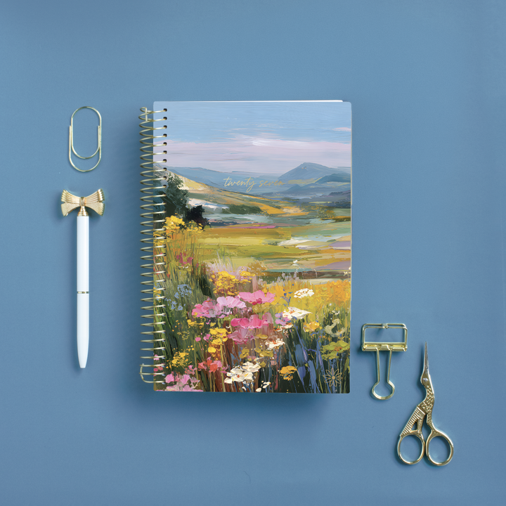 A spiral-bound notebook with a colorful wildflower and mountain landscape cover sits on a blue background, styled with gold paperclips, a white pen with a gold bow detail, and gold scissors.